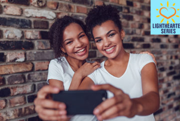 Two happy young girls in white v neck t shirts take photo of them together on a smartphone, against a brick wall