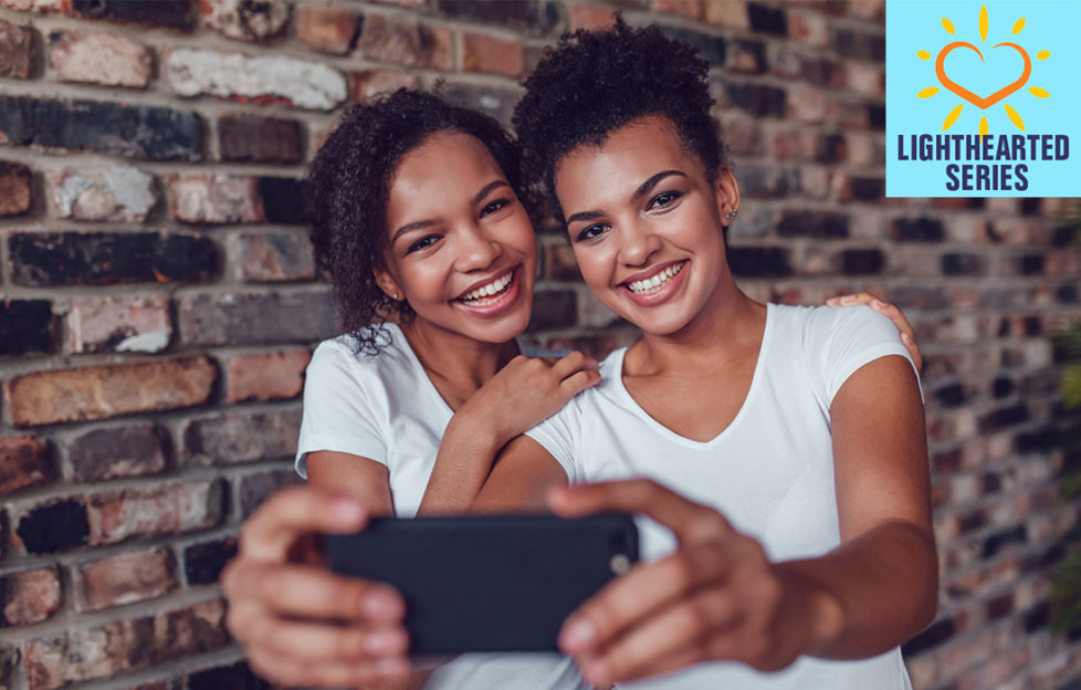 Two happy young girls in white v neck t shirts take photo of them together on a smartphone, against a brick wall