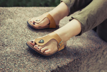 Woman's feet in yellow stylish summer sandals with dark nail polish, in green summer pants sitting on the wall;