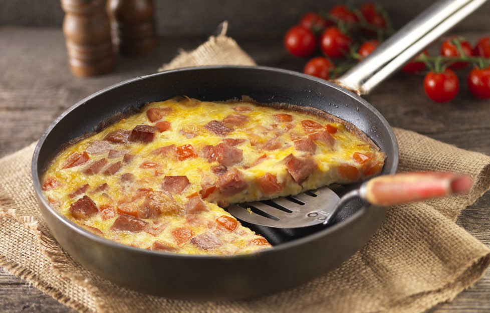 Frying pan on piece of sacking, one slice cut from golden Spam frittata, deep red tomatoes behind