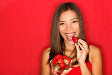 Woman eating strawberries happy. Pretty girl eating healthy snack on red background. Attractive Asian Caucasian female model joyful.