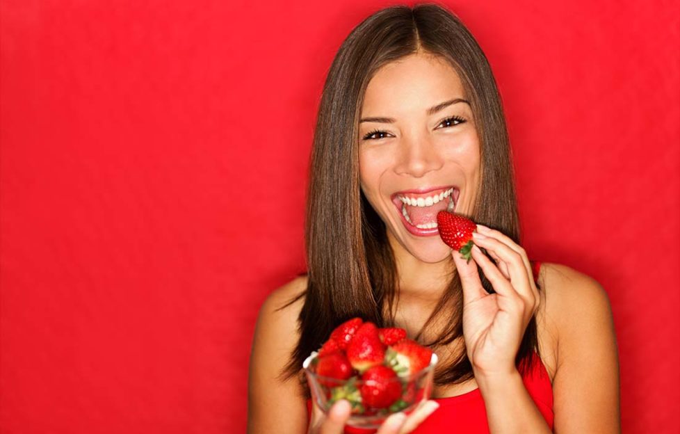 Woman eating strawberries happy. Pretty girl eating healthy snack on red background. Attractive Asian Caucasian female model joyful.