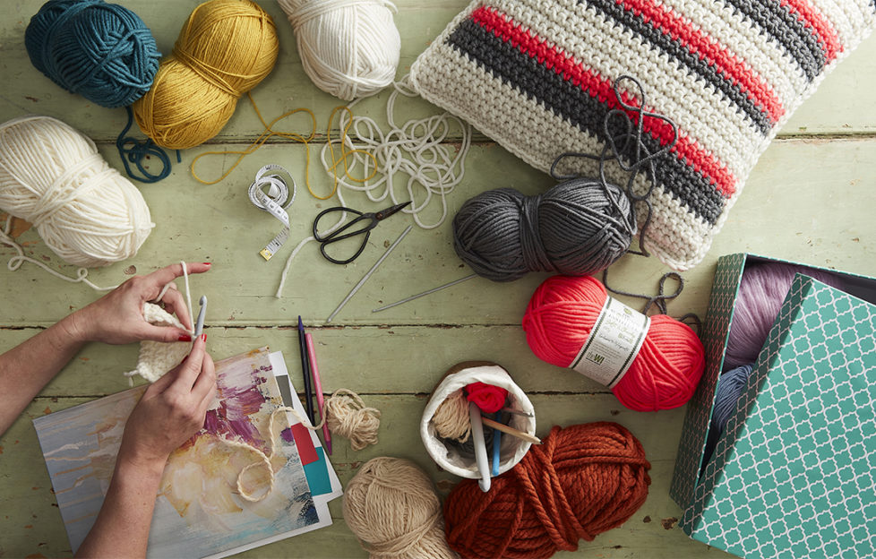 Crafter's desk with balls of yarn, scissors, pot of crochet hooks, crocheted cushion and woman's hands crocheting. All ready to get started with some mindfulness crafts.