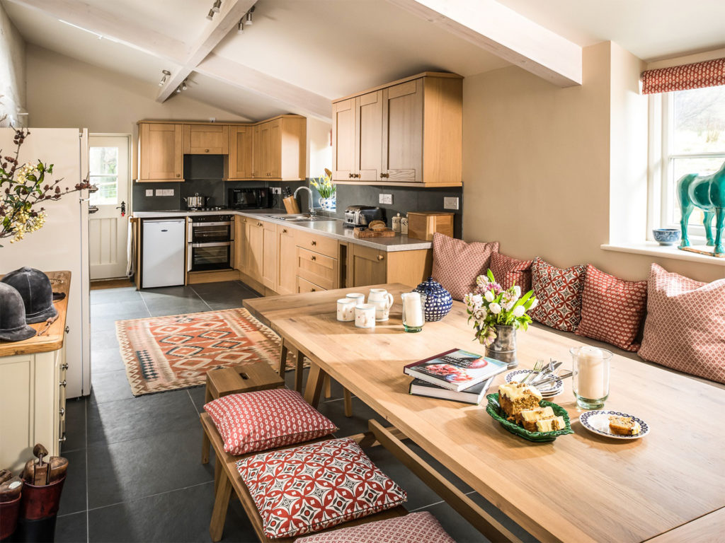 Large open plan kitchen diner with black slate floor, wooden cupboards and long table with patterned cushions on benches