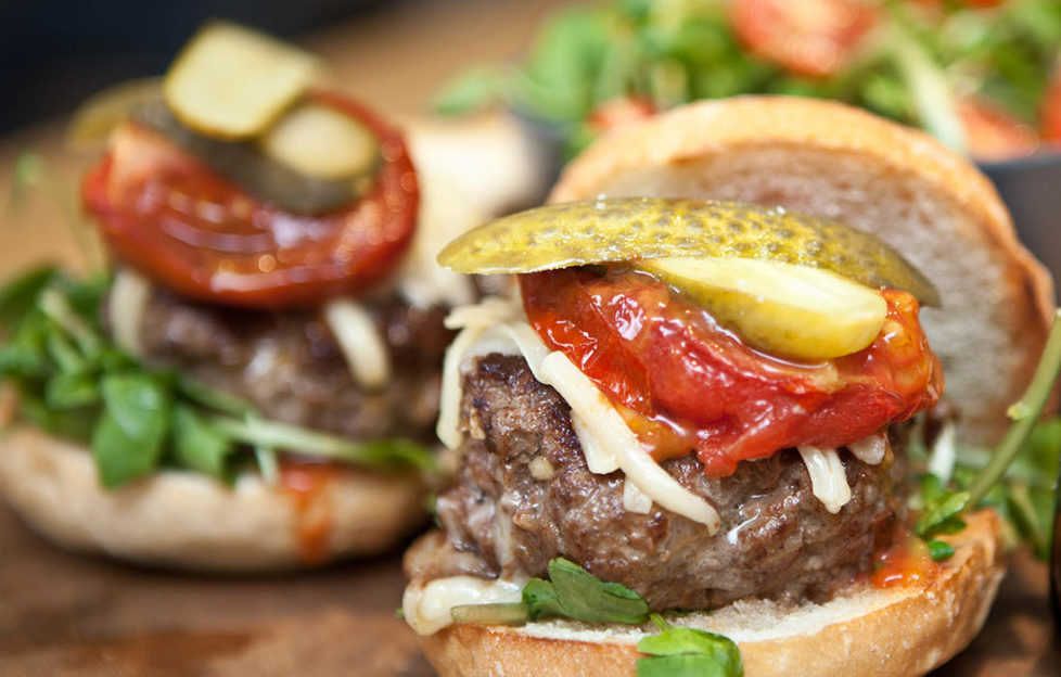 Close up of large, juicy home made burger with umami flavours -with sunblush tomato, cheese, watercress and sliced gherkin