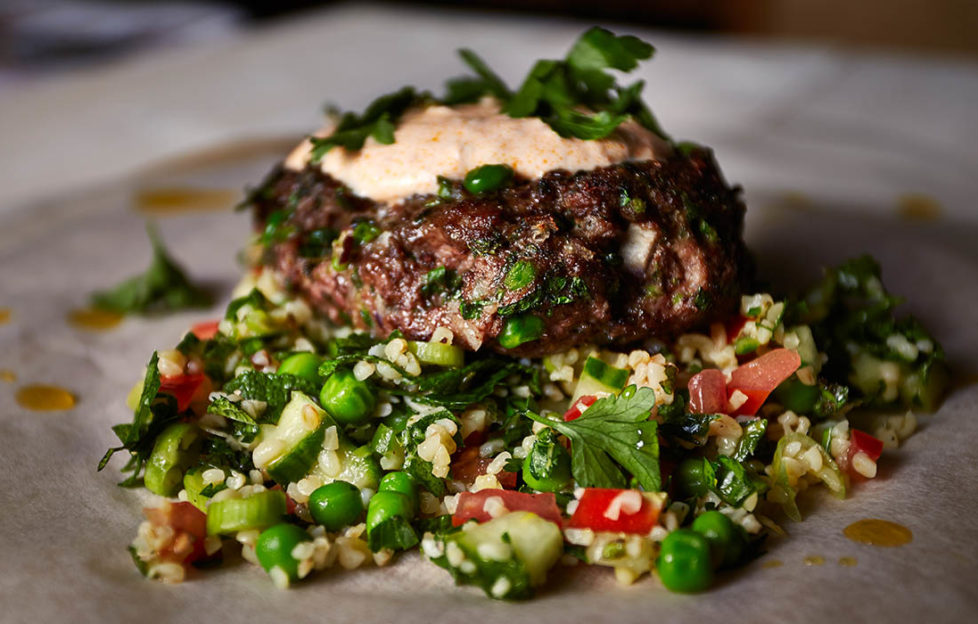 Juicy lamb kofta burger topped with rose yogurt and herbs, sitting on pile of tabbouleh, grains, chopped herbs and salad veg.