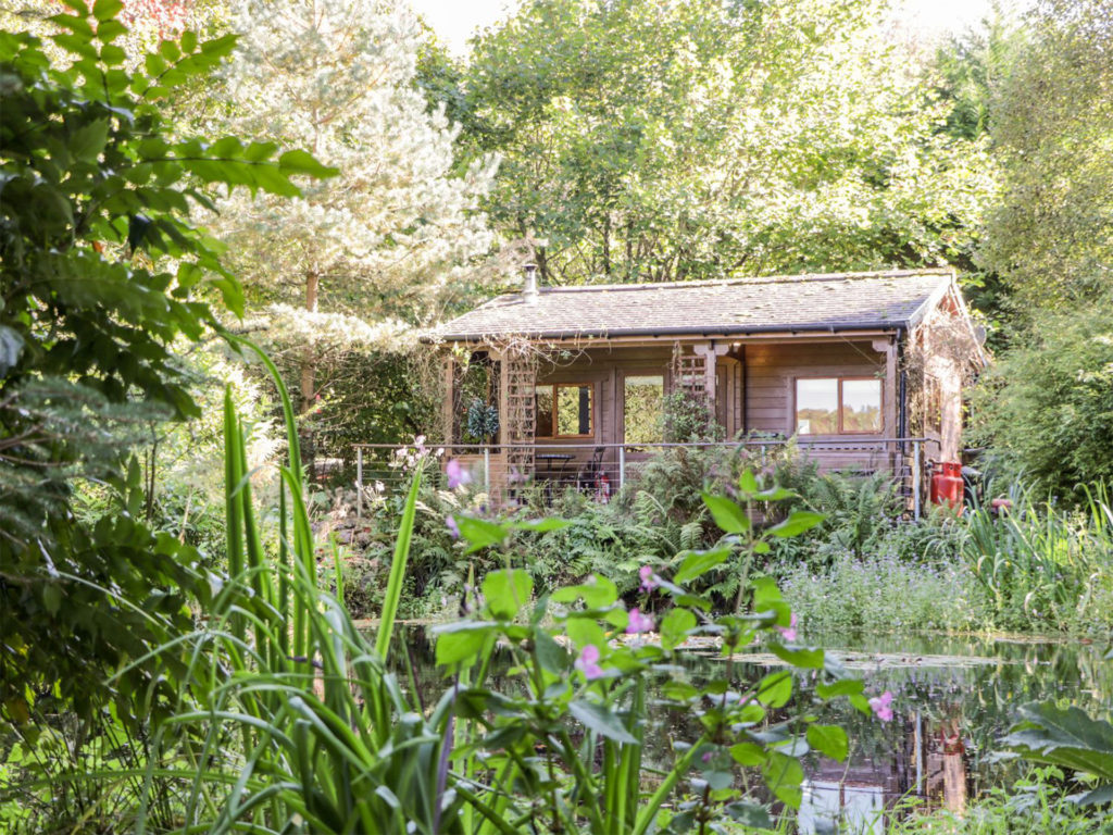 Log cabin set in trees and luxuriant garden with pond