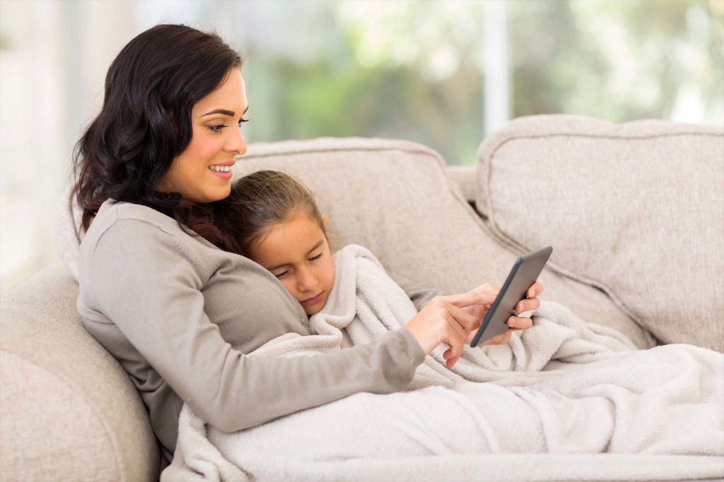 mother sitting on the couch with her sleeping daughter and using tablet computer