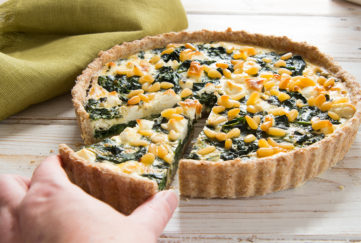 Feta quiche containing spinach and pine nuts, hand taking one cut slice