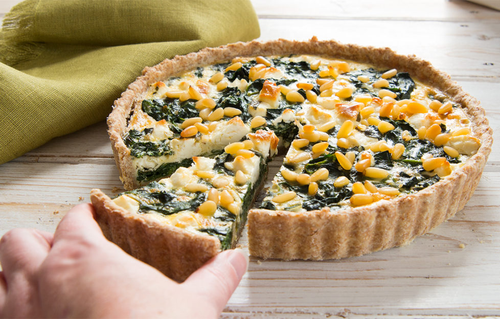 Feta quiche containing spinach and pine nuts, hand taking one cut slice