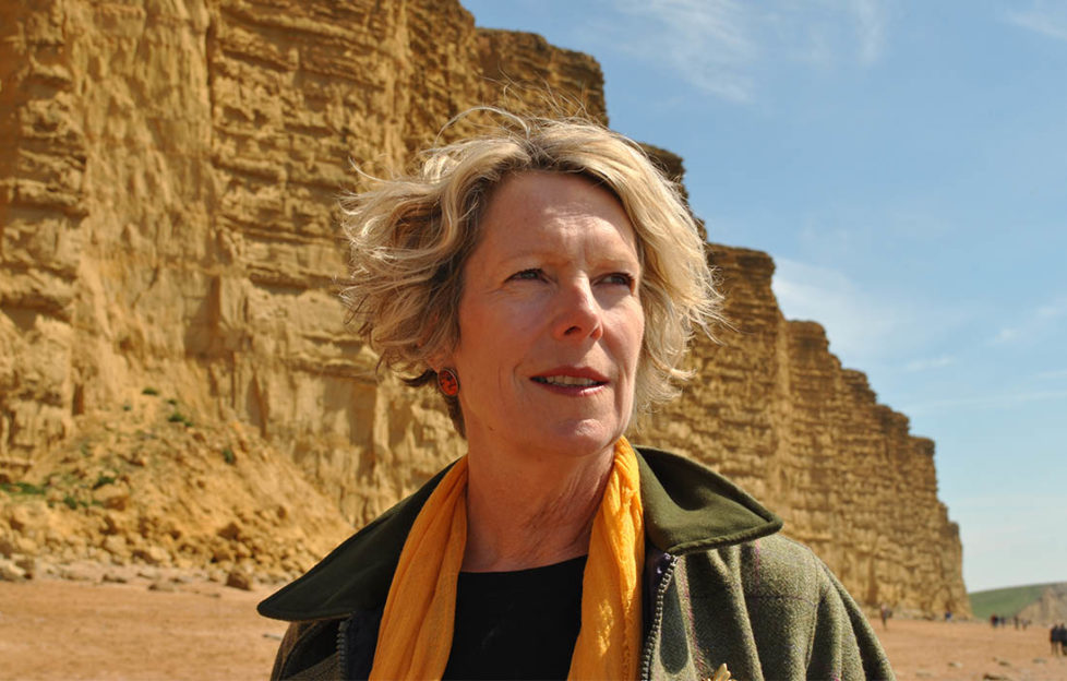 Author Rosanna Ley, blonde middle aged woman in rich yellow scarf and green walking jacket standing by yellow cliffs