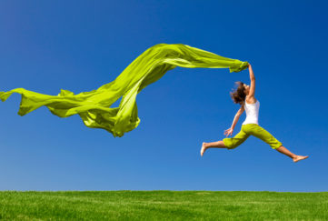 Beautiful young woman jumping on a green meadow with a coloured tissue;
