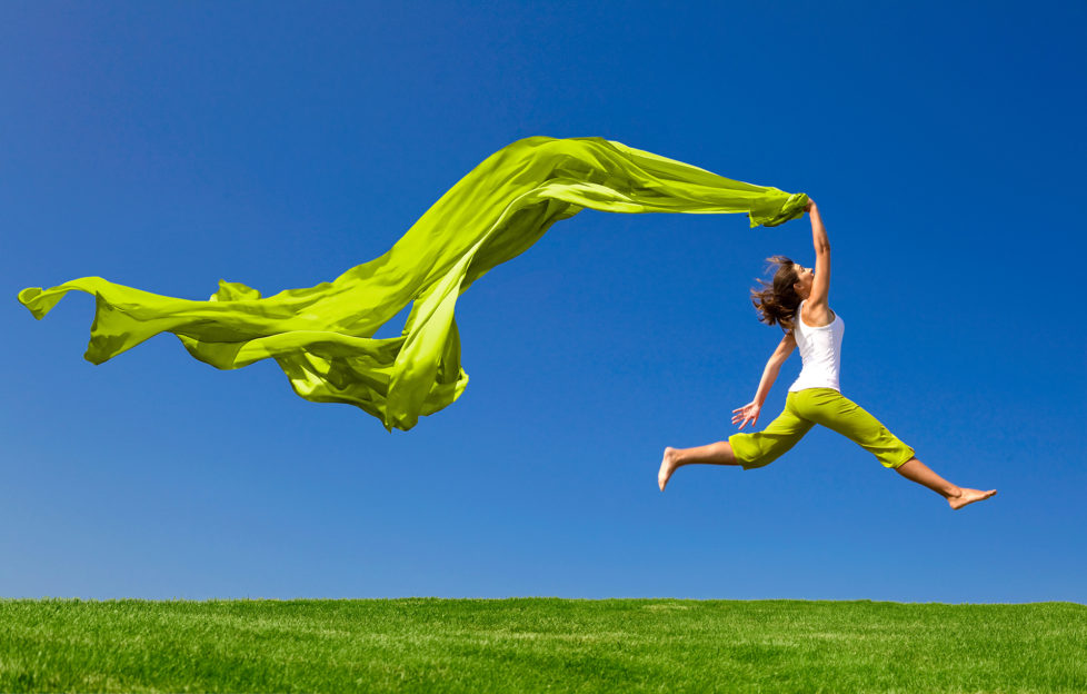 Beautiful young woman jumping on a green meadow with a coloured tissue;