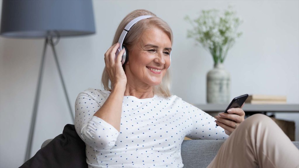 Smiling mature woman wear wireless headphone hold smartphone looking at phone screen using mobile player app listening online music, learning foreign language, watching video relaxing on sofa at home