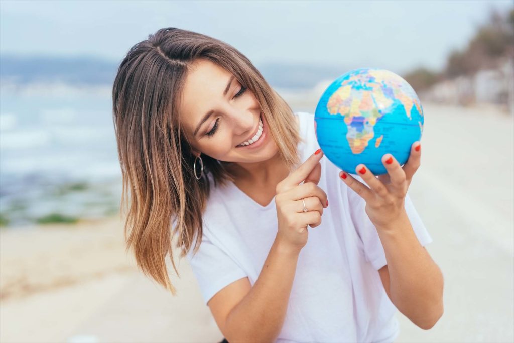Smiling pretty young woman pointing to her holiday destination on a handheld world globe as she stands on a seafront promenade