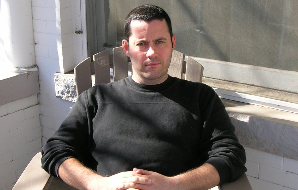 Crime writer Adrian McKinty sitting outside a house on a wooden chair, wearing plain black jumper
