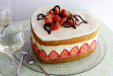 Heart shaped strawberry cake topped with white marzipan, strawberries and chocolate hearts. Between the sponge layers is a deep layer of set custard and a row of sliced strawberries arranged vertically.