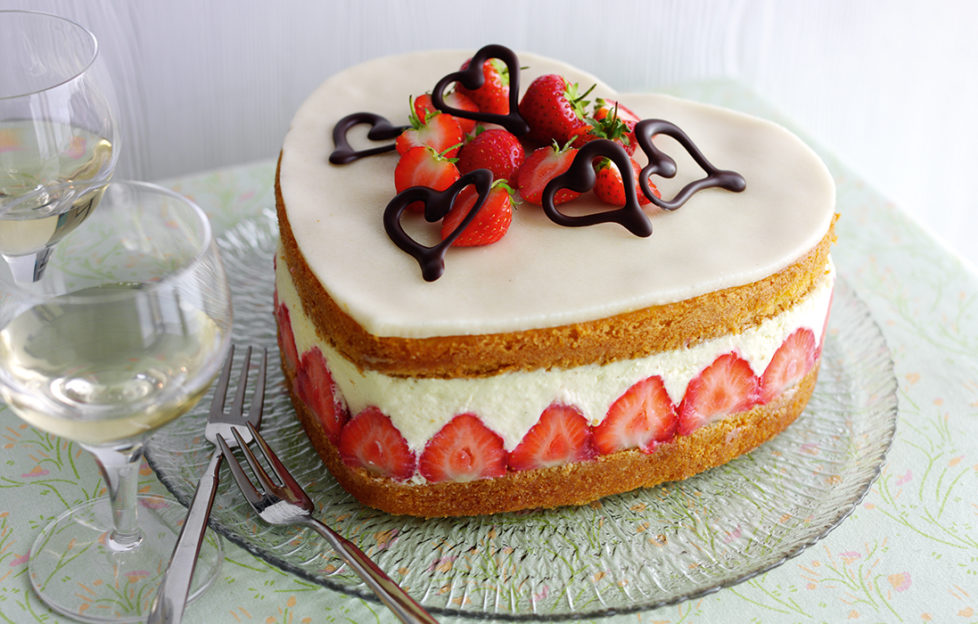 Heart shaped strawberry cake topped with white marzipan, strawberries and chocolate hearts. Between the sponge layers is a deep layer of set custard and a row of sliced strawberries arranged vertically.