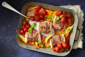 Brightly coloured tomatoes, fresh basil, pieces of fish wrapped in parma ham and other vegetables in a baking tray