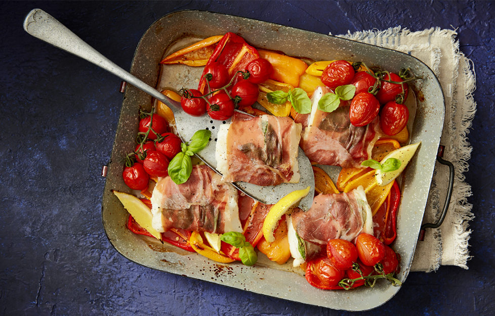 Brightly coloured tomatoes, fresh basil, pieces of fish wrapped in parma ham and other vegetables in a baking tray