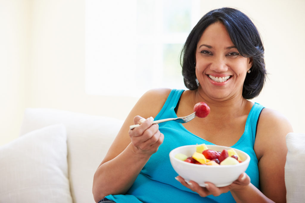 Overweight Woman Sitting On Sofa Eating Bowl Of Fresh Fruit; 