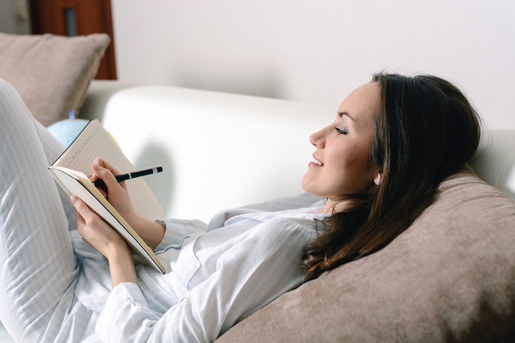 Satisfied and happy smiling girl lying on the sofa in the room and writes a journal of your dreams, plans, goals, experiences, ideas, lived emotions and feelings.;
