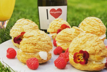 Plate of cream puffs on a plate outdoors with raspberries, flowers and I Heart Prosecco bottle