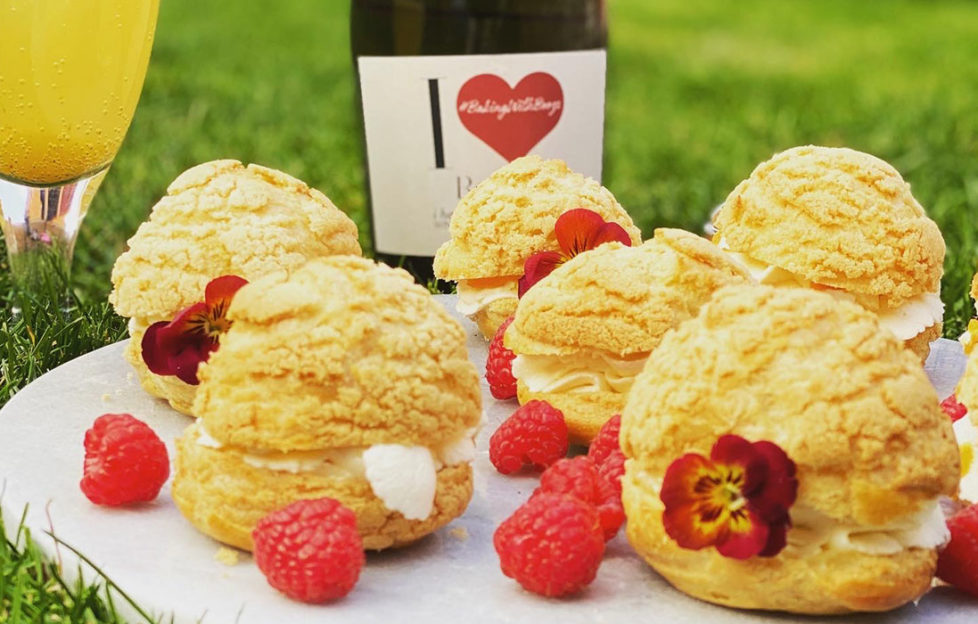 Plate of cream puffs on a plate outdoors with raspberries, flowers and I Heart Prosecco bottle