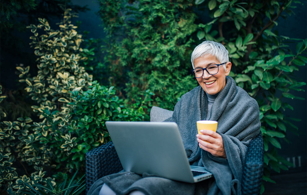 Mature woman in garden, wrapped in grey blanket, holding cup of tea, using laptop