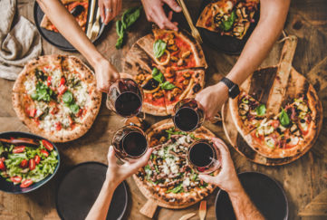Pizzas on table, 4 people reaching across to clink glasses of red wine, seen from above