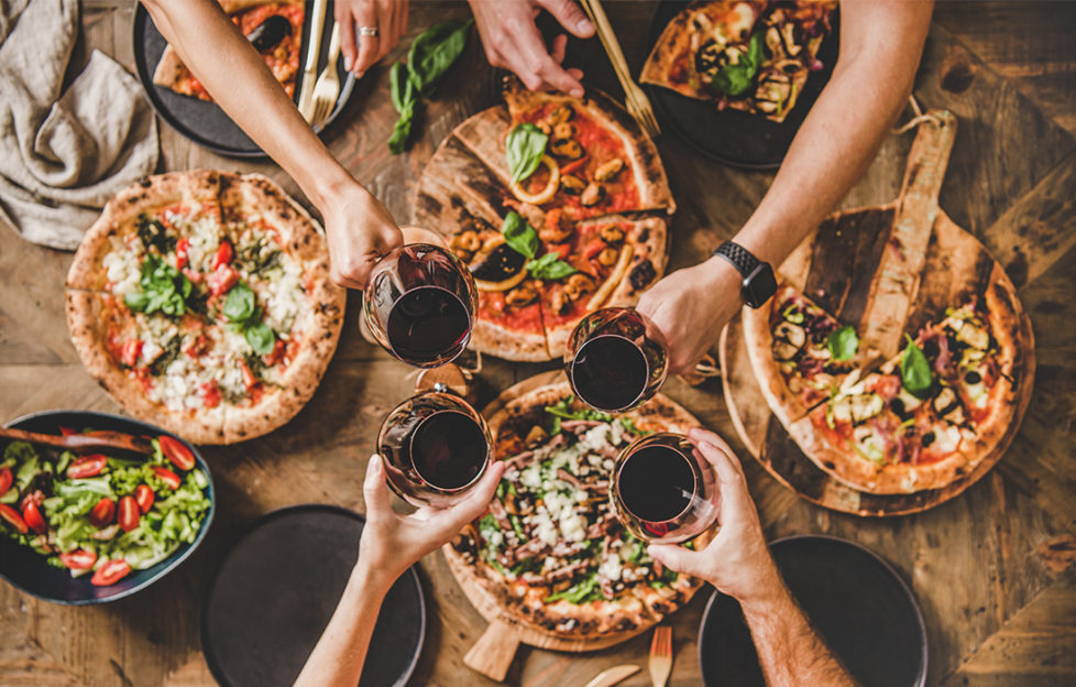 Pizzas on table, 4 people reaching across to clink glasses of red wine, seen from above