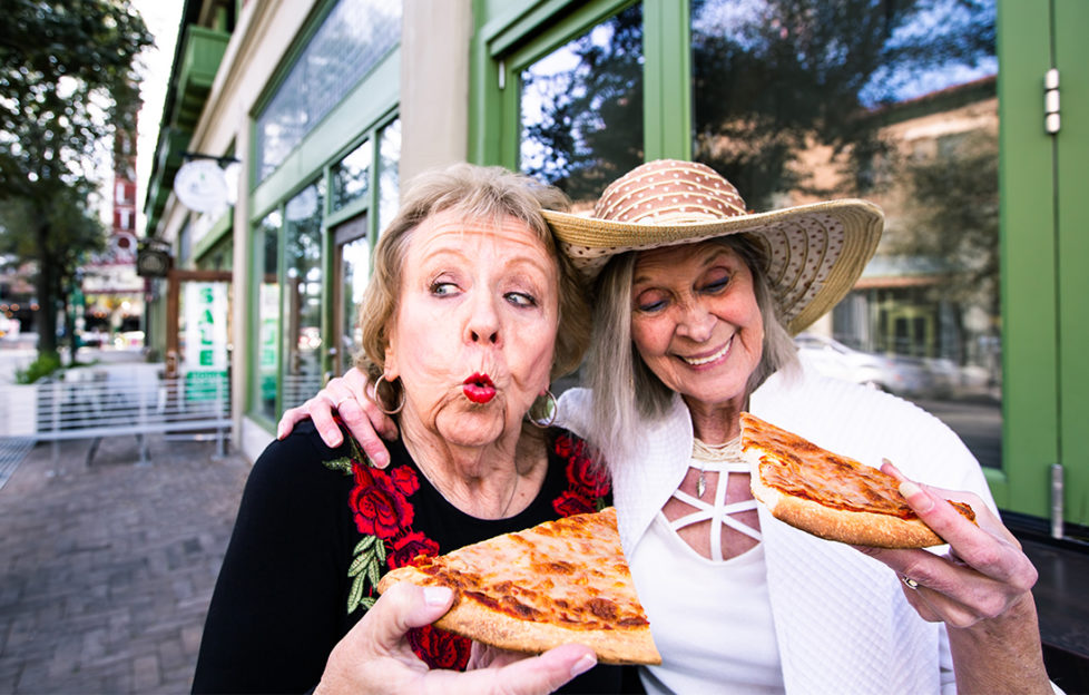 2 mature women eating street food pizza, pulling funny faces, guilty pleasure