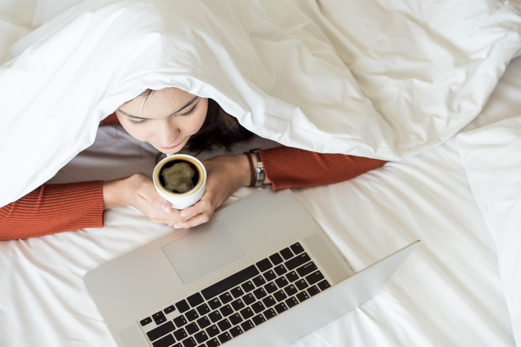 Woman under duvet with mug of coffee, working on laptop, seen from above