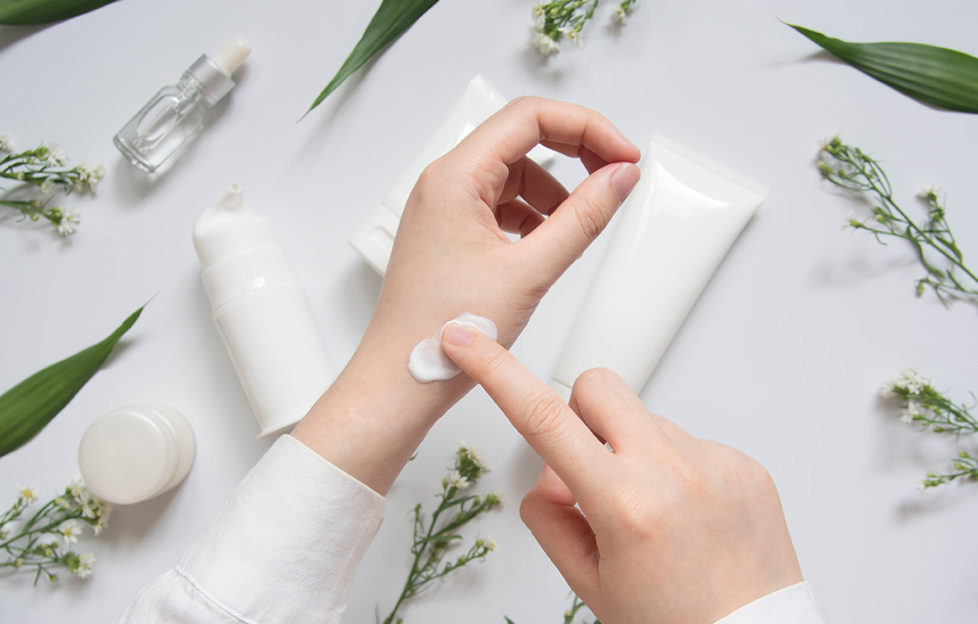 Testing natural ingredients. Woman applying cream to her wrist, various plain tubes and pots on table, sprigs of herbs laid out around