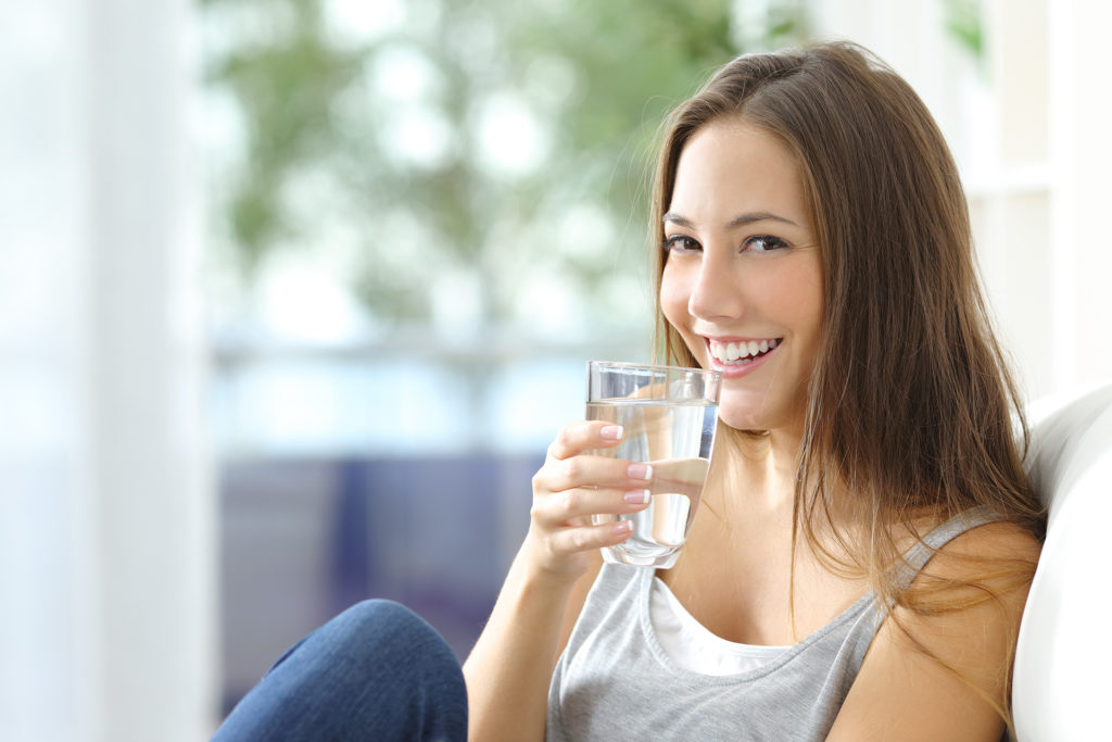 Girl drinking water sitting on a couch at home and looking at camera; 