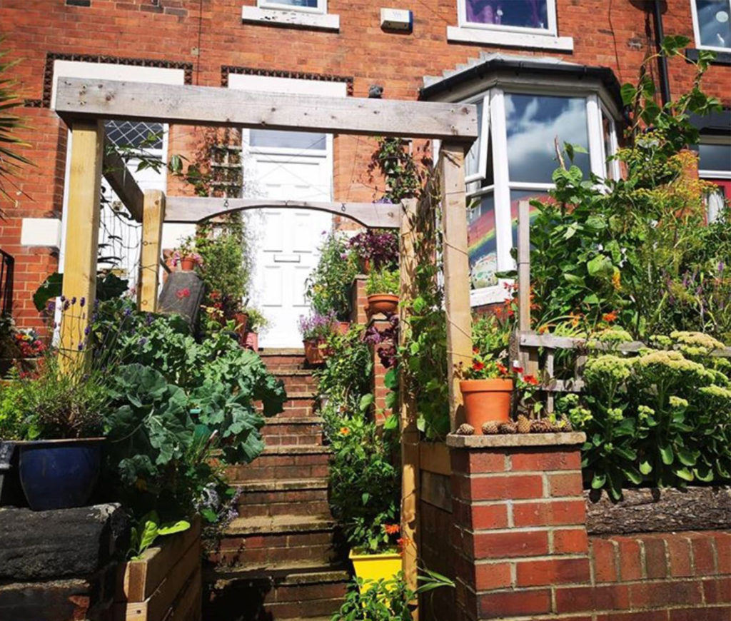 View from bottom of front steps. Red brick terrace with wooden frames, containers and lush plants in small space in front of house