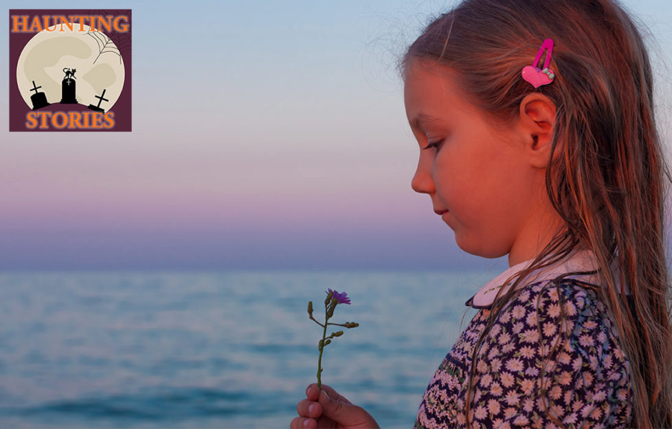Girl aged around 8 stands by sea at dusk, looking at wild flower, she has long hair and a traditional summer dress with floral print and white collar