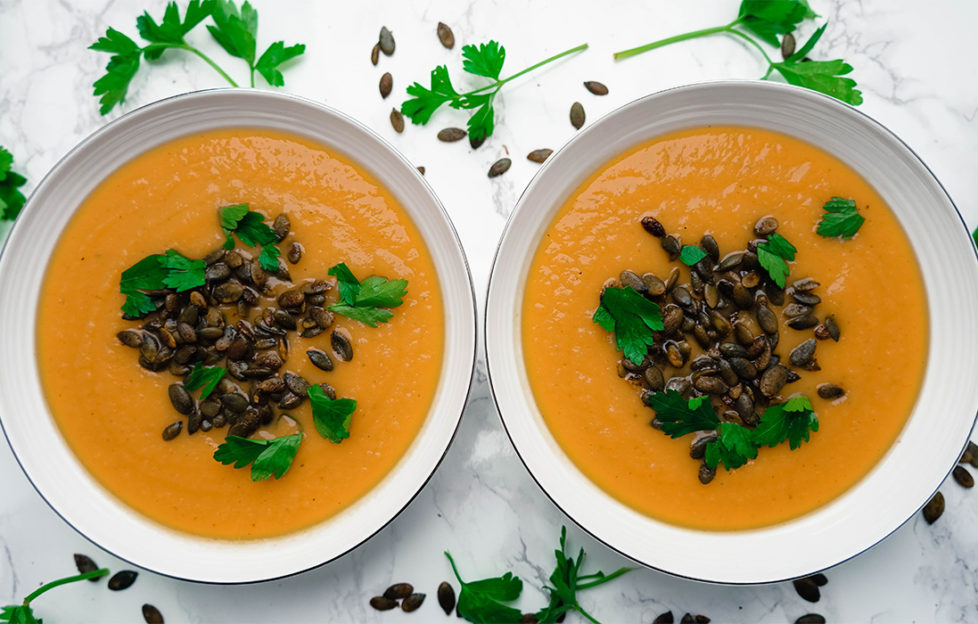 2 bowls of orange soup topped with toasted seeds, flat leaf parsley sprinkled around