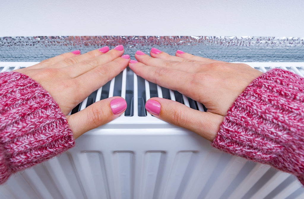 Woman warming hands wearing warm clothes near heating radiator. Heating season at home, concept.;