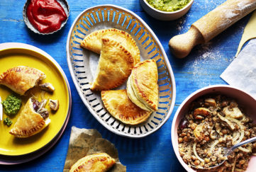Half moon shaped mini pasties in an oval dish, turquoise background,dishes of avocado salsa, ketchup and lentil filling to side