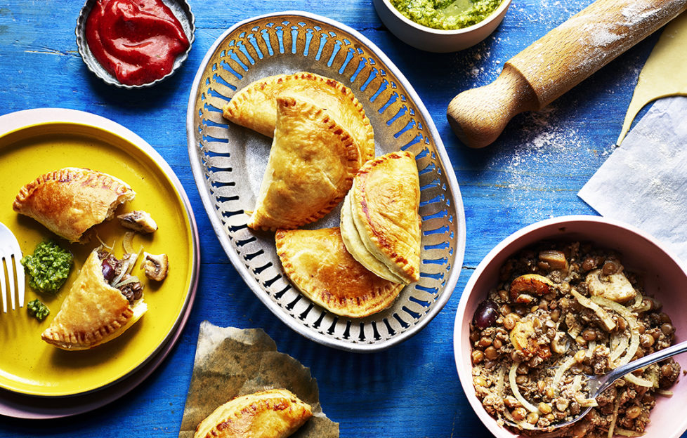 Half moon shaped mini pasties in an oval dish, turquoise background,dishes of avocado salsa, ketchup and lentil filling to side