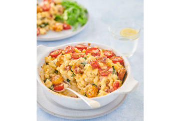 A bowl of mac 'n cheese topped with halved cherry tomatoes and with a fork resting on the side sits on a white table cloth. There's a glass of lemon water and another plate of food out of focus behind it.