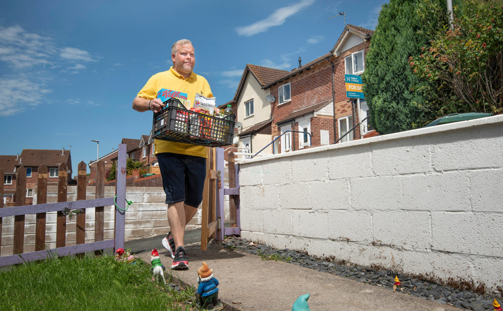 Man walks up garden path past garden gnomes with crate of groceries
