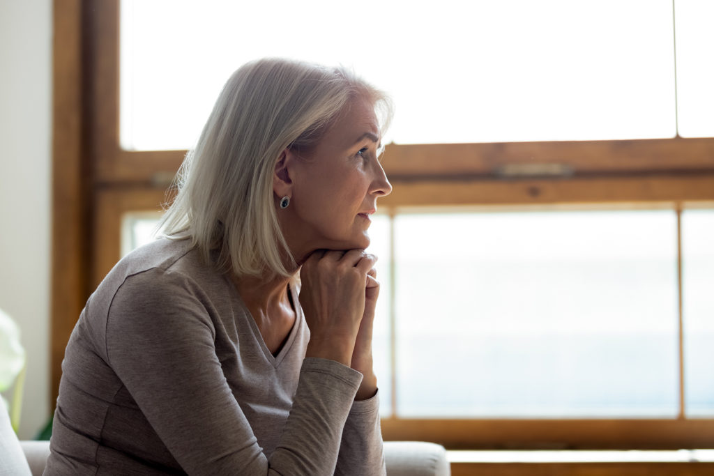 Side view of sad thoughtful middle-aged mature woman sit on couch at home look in window distance mourning, upset pensive senior female lost in thoughts thinking or pondering over past; 