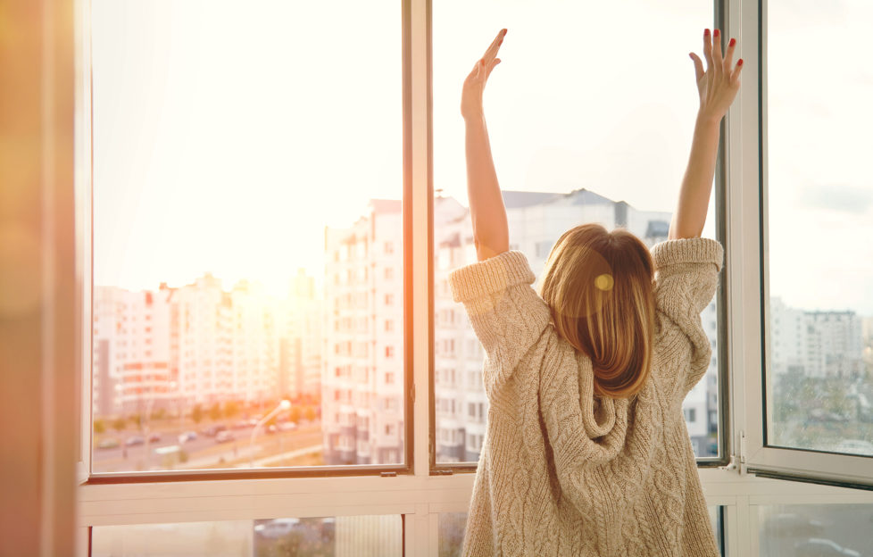Woman near window raising hands facing the sunrise at morning;