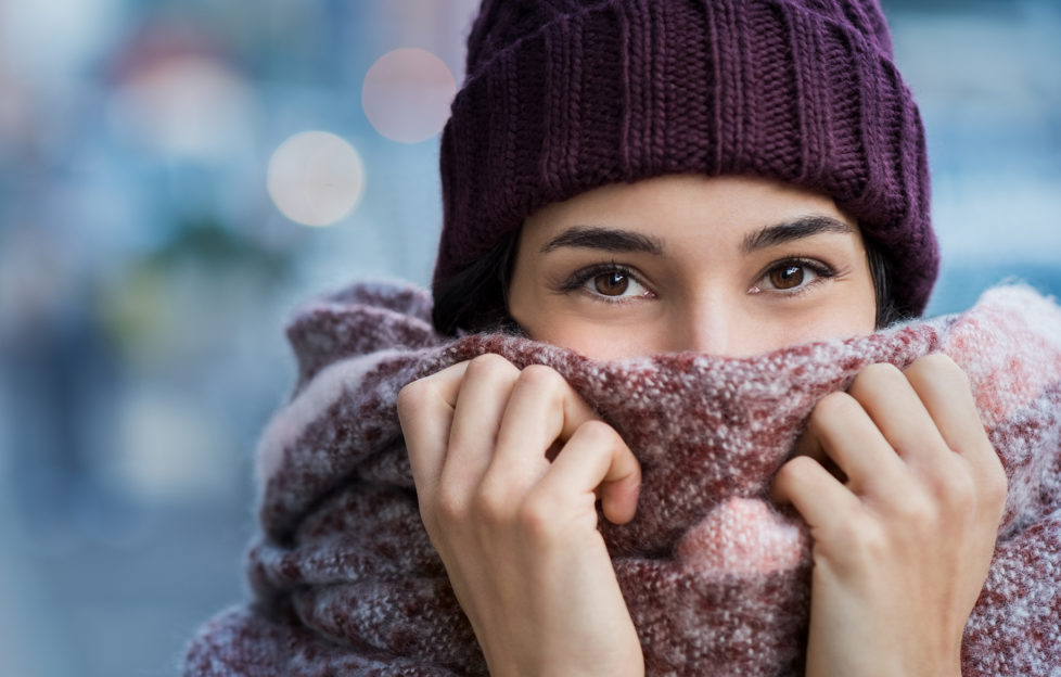 Winter portrait of young beautiful woman covering face with woolen scarf. Closeup of happy girl feeling cold outdoor in the city. Young woman holding scarf and looking at camera.