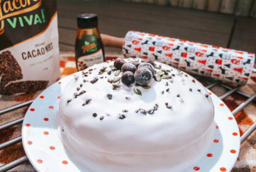 Christmas cake covered in royal icing, decorated with nuts, cocoa nibs and blueberries, Christmas patterned rolling pin and pack of Yacon Viva cocoa nibs behindbehind