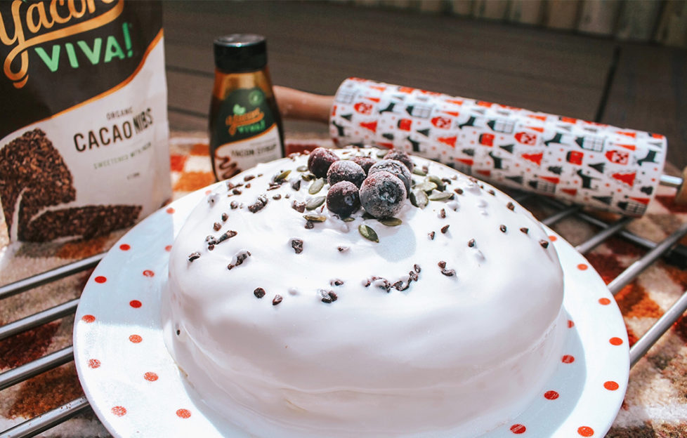 Christmas cake covered in royal icing, decorated with nuts, cocoa nibs and blueberries, Christmas patterned rolling pin and pack of Yacon Viva cocoa nibs behindbehind