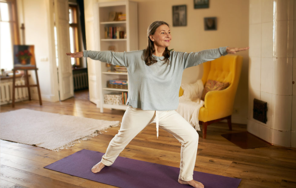 Full length shot of happy energetic mature woman in casual clothes exercising at home because of social distancing, practicing yoga on mat, standing in warrior ii pose. Age, wellness and health;