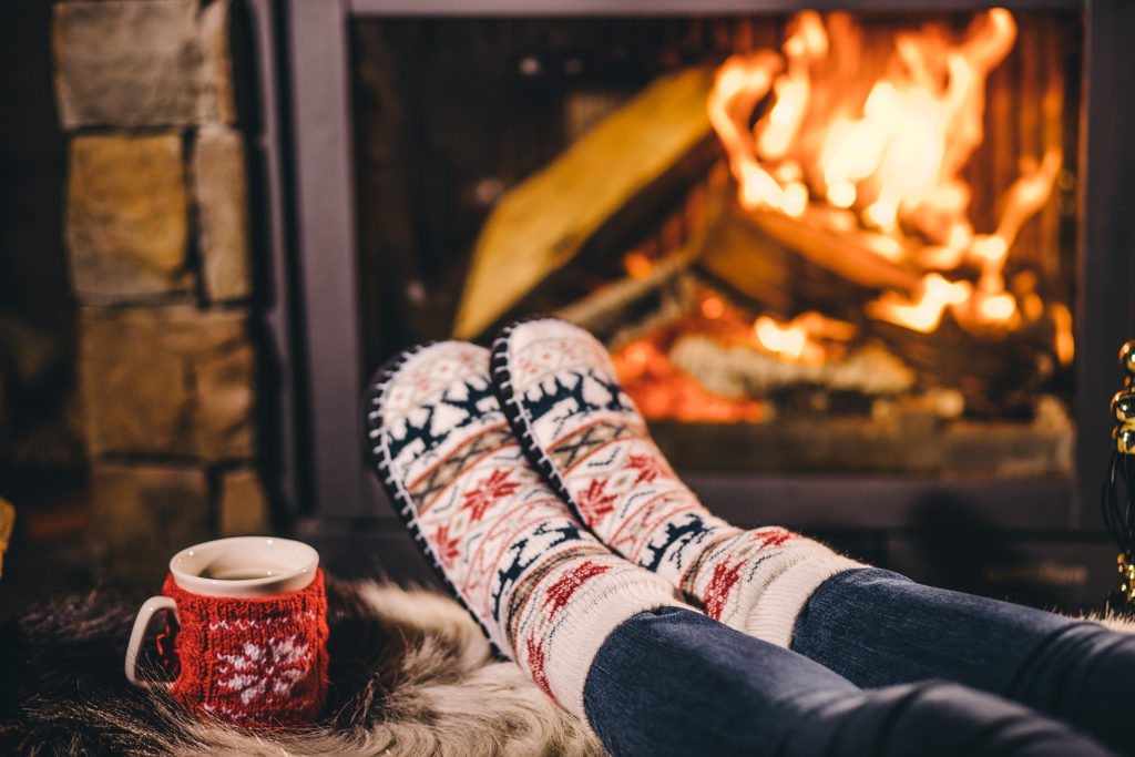 Feet in woollen socks by the Christmas fireplace. Woman relaxes by warm fire with a cup of hot drink and warming up her feet in woollen socks. Close up. Winter and Christmas holidays concept. ; 